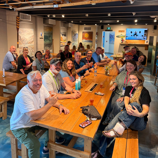 A group of people sitting around a large wooden table in a museum or exhibition space, holding drinks and smiling and talking.