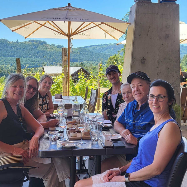 A group of seven people sitting together at an outdoor dining table on a patio, with a large umbrella, vineyard rows, and mountains in the background.
